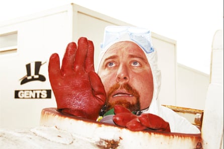 A man in a white hazmat suit and red gloves sitting in a dirty white barrel, with his head and shoulders emerging. In the background is a portable loo with a sign reading “GENTS”.