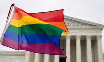 Rainbow flag waves in wind under cloudy sky in front of peaked, columned, white-stone building.