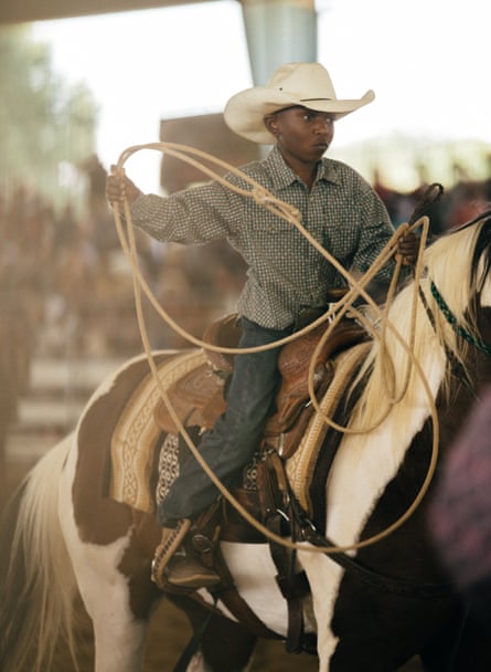 A young Black boy, middle school age, wearing a white cowboy hat, jeans shirt, jeans an cowboy boots, holding a lasso almost as big as he is, on a brown and white horse.