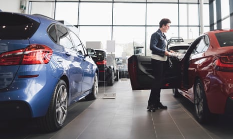 anonymous woman looking inside red saloon car with open front door in a modern glass showroom; it is parked by a metallic-blue hatchback but no brand-names are visible