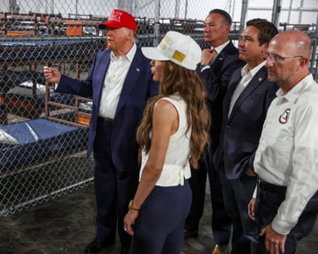 Four men and one woman stand on other side of chain-link fence from rows of bunks.