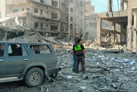 One man with his arm around the other’s shoulders as they look at the rubble of destroyed buildings