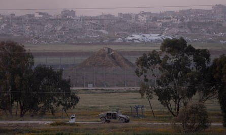 An army vehicle is driven along a track across grassland in front of heaps of earth and high wire perimeter fencing; there are destroyed buildings and a town in the distance in the background