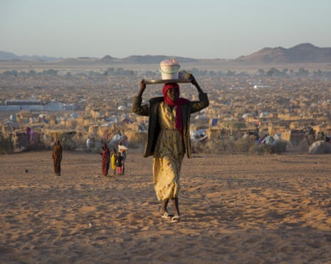 A woman carries a tray and bucket on her head as she walks across a stretch of empty ground, with the tents and grass huts of the Tawila displacement camp stretching into the distance behind her