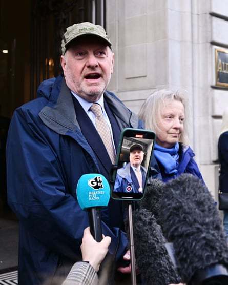 Alan Bates, in a baseball cap and suit and tie under a raincoat, stands outside a building next to a woman and speaks into press microphones