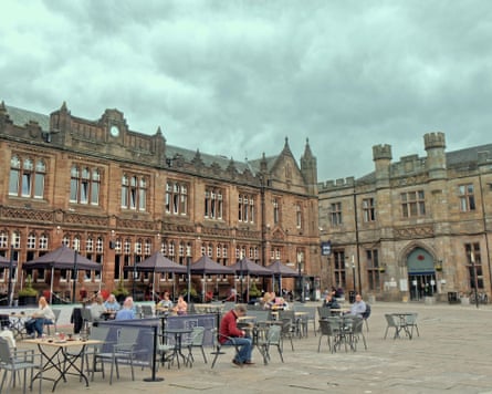 People drinking outdoors in a square surrounded by stone buildings under grey skies