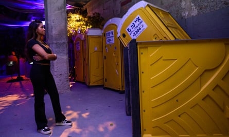 A young woman stands with her arms folded looking at a row of yellow portable toilets, some of which have fully or partly fallen over