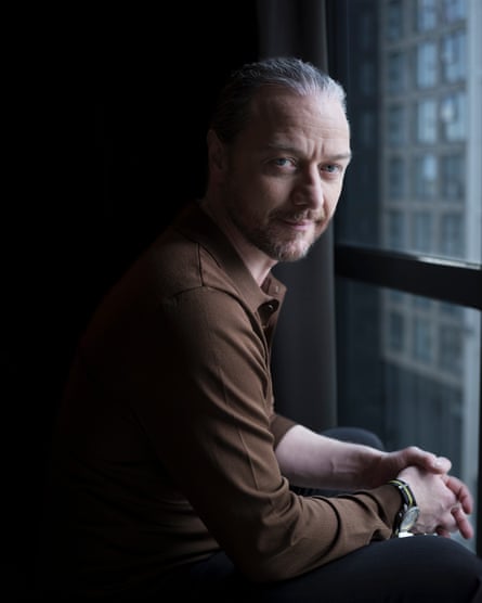 James McAvoy photographed in Glasgow, March 2026. He sits by a floor-to-ceiling window, the light illuminating his face in an otherwise dark room