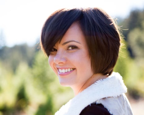 Woman with straight hair grins as she looks over her shoulder with sunshine and greenery behind her