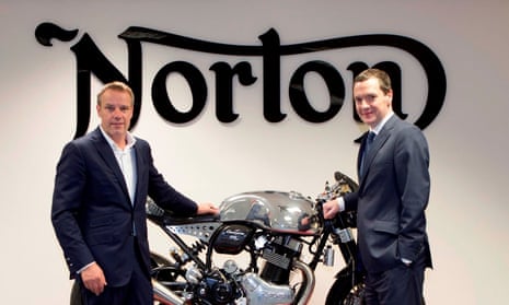 Two men stand either side of a motorcycle manufactured by Norton, whose logo is in the background