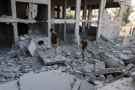 Two residents walk over rubble in front of a bombed out building.