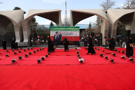 Women dressed in black dresses and black hijabs walk over a red carpet that has rows of stools lined up on it in front of a smiling photo of Ayatollah Ali Khamenei against a red, white and red backdrop.