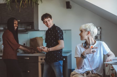 A young woman at a laptop looks round at her roommates, who are making breakfast behind her
