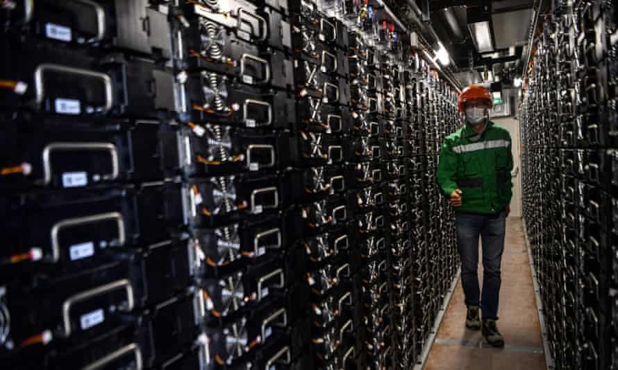 Rows and rows of small black containers with handles stacked up inside a building, with a man in a coronavirus face mask walking in the corridor between them