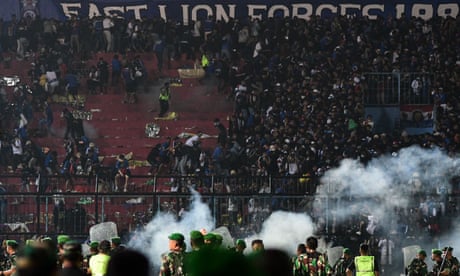 Fans berkerumun di sisi tribun di stadion, dengan polisi dan gas air mata terlihat di latar depan.