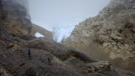 Two men standing at the moot of a section of brown mountain terrain with a small white glacier in the background