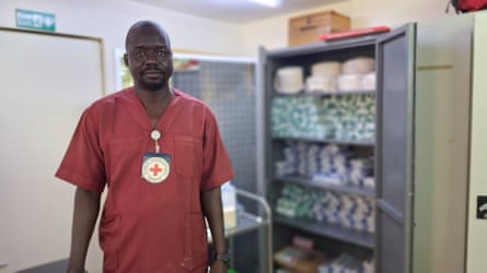 A man in red medical scrubs stands in front of an open cabinet containing medical supplies