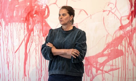 Tracey Emin photographed in front of one of her pictures; she wears dark clothing which contrasts with the red, pink, white and pale cream of the painting. She stands with her arms folded and looking to one side; her dark hair is tied back.