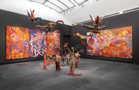 A gallery space with black walls and a dark grey floor, with a series of colourful paintings on the walls, and a cluster of woven sculptures of figures and creatures in the centre of the room, some installed on the floor and several suspended from the ceiling.