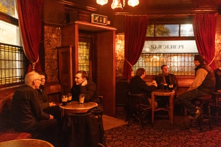 The Palm Tree in Mile End, London, at night, showing two tables of people in a traditional pub interior at night, talking with pints in front of them. There are velvet curtains at the windows, patterned carpets and old-fashioned overhead lighting.