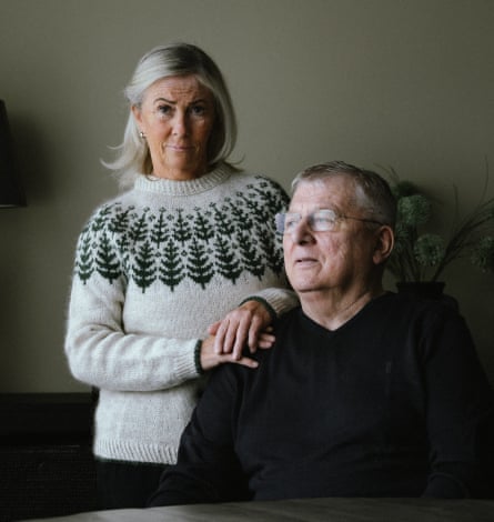 A woman with a grey bob wearing a cream sweater with pine trees on it, standing next to a man sitting by a table wearing a black jumper