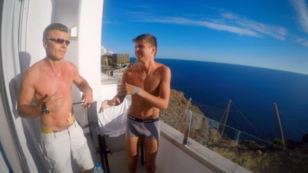A bare-chested Gunnar Hall Jensen and his teenage son on a hotel balcony laughing