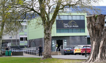 Ysgol Dyffryn Aman: view of the frontage of a modern school building with light green panelling and a sign with the school’s name. Vehicles including a police car are parked in front and police tape is cordoning off the entrance