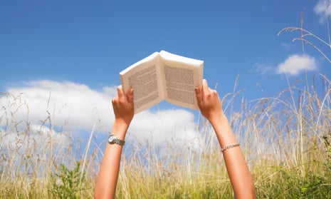 A person's arms are extended towards the sky holding open a book. The person is lying in a field of grass under a blue sky with a few clouds