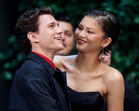 Tom Holland and Zendaya smile together at a photo call, he in a black suit, she in a strapless dress
