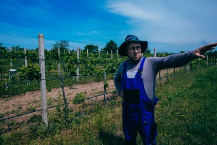 Heorhii Molchanov, a winemaker, dressed in a hat and blue overalls pointing to the right as he shows the family vineyards. On the very left, the unexploded Russian rocket stuck in the ground.