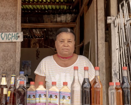 A woman stands in a wooden hut behind a row of bottled drinks. A wooden sign says ‘viche’ next to her