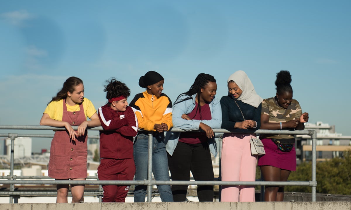 Image is from the film 'Rocks' (2020). A group of six teenager girls lean against a railing and chat to one another. Behind them is the London skyline.