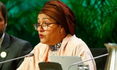 Amina Mohammed, seen here at the Cop15 biodiversity summit in 2022: she is in her early 60s and wears a brown head-covering and pale, shiny robe over a patterned top; she is seated at a microphone as she addresses the conference and is wearing glasses and looks serious.