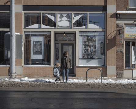 A child plays in the snow outside of a storefront and holds her mothers hand