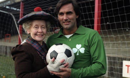 Gudrun Ure as Super Gran with George Best: she is wearing a large floppy tartan hat with red pompom, and Best wears a green sports top with white shamrock symbol. They are standing together holding a football.
