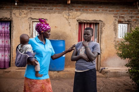 A tall African woman outside a brick-built house holding a baby in one arm touches the arm of a girl standing next to her