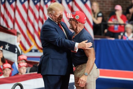 Man in blue T-shirt and khakis hugs Trump, wearing blue suit in front of row of American flags