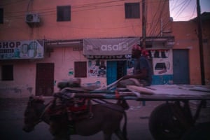 A man sits on a cart led by a donkey in a city street