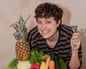 Lucy by a pile of fruit and veg, holding up a peeler and smiling