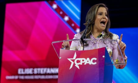 White woman, long brown hair, mouth wide as if speaking and pointing one finger, standing at pink lectern with CPAC logo.