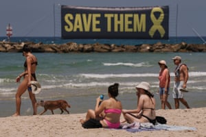 People sit or walk on a sandy, sunny beach. In the distance there is a large banner with the words ‘Save them’