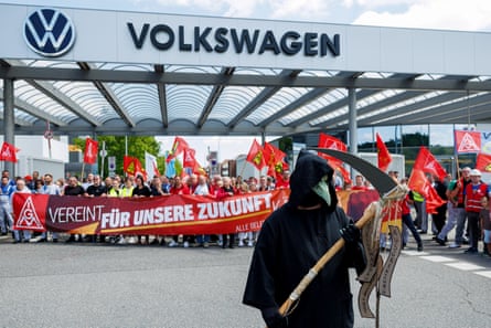 A protester wearing a black Grim Reaper costume with hood and beaked mask, and carrying a sickle, stands in front of the Zwickau plant’s entrance and Volkswagen sign, where a large group of employees are holding flags and banners.