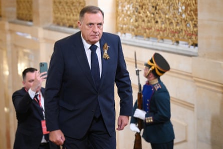Milorad Dodik arrives for a gala concert in Moscow. He is heavily built and wears a dark suit with a loop of striped ribbon in his buttonhole. He is walking up stairs in a grand building with gold decoration.