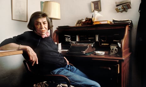 Patricia Highsmith in jeans, with short hair, leaning against a shelf while sitting at her old wooden desk with a typewriter