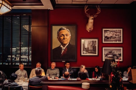 people sit at a long table in a restaurant, a large painting of a man on the wall above them