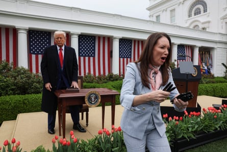 Donald Trump stands at a desk on a platform in the White House garden while a woman hands out pens to people off camera