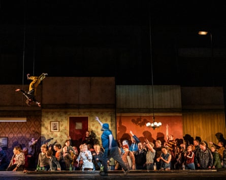 A heavyset man on stage in front of a crowd of amazed-looking people points at a man suspended upside down in the air