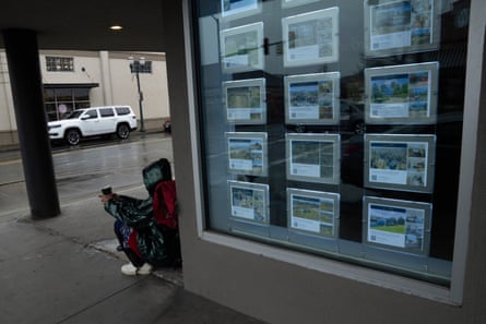 person sits on the ground next to a shop with pictures of houses for sale in the window