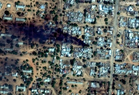 Aerial view of a town with a grid of roads and compounds, dotted with trees and a plume of black smoke