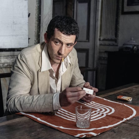 Josh O’Connor in a beige suit, smoking and sitting at a low wooden table